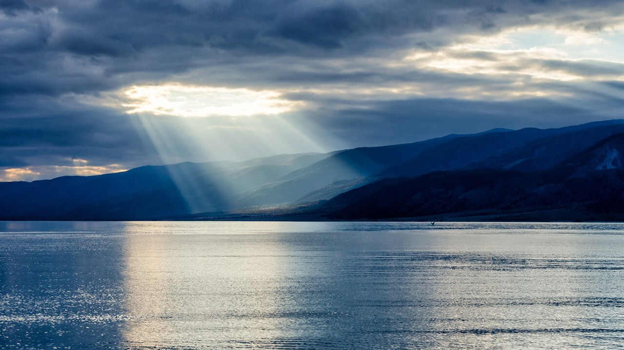Sunbeams warmly illuminate a body of water with majestic mountains in the background, filling the onlooker's heart with gratitude for this breathtaking display at this specific latitude.