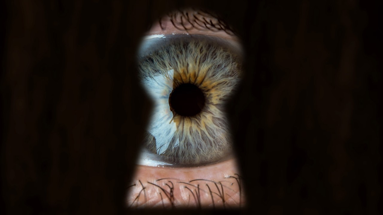 A person's eye, filled with fear, peeking out of a keyhole.